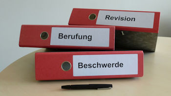Die Aufnahme zeigt im Hintergrund drei liegende rote Ordner mit den Aufschriften "Revision, Berufung und Beschwerde". Im Vordergrund ein liegender Kugelschreiber.